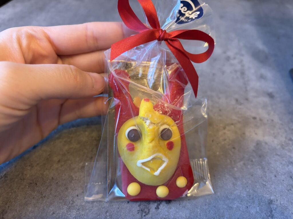 透明の袋に入った赤いチョコレートの人形。黄色い丸いお面に茶色い目、赤いほっぺたが描かれており、赤いリボンで袋の口が結ばれている。手のひらに乗せて持っている様子。