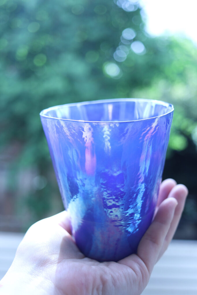 ブラーノ島のお土産に購入した青紫色の手吹きガラスのコップ。ガラス工房Artistico Bombon製。自然光の中で手に持って撮影。