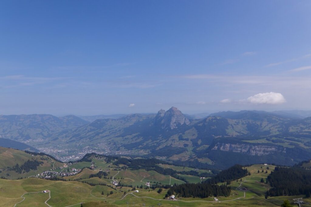 クリンゲンシュトックから見下ろす大パノラマ。緑の牧草地と山あいの町、中央にミューテン山の尖った峰